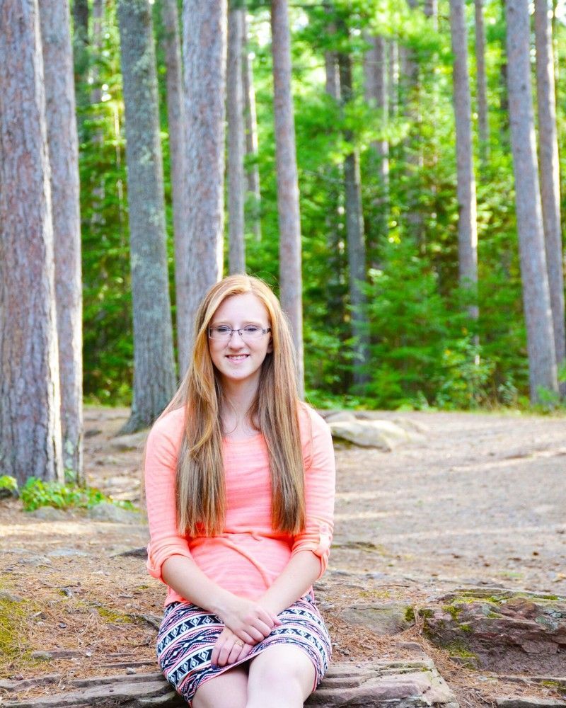Woman in a coral top and patterned pants sitting on a rock in a forest, smiling at the camera.