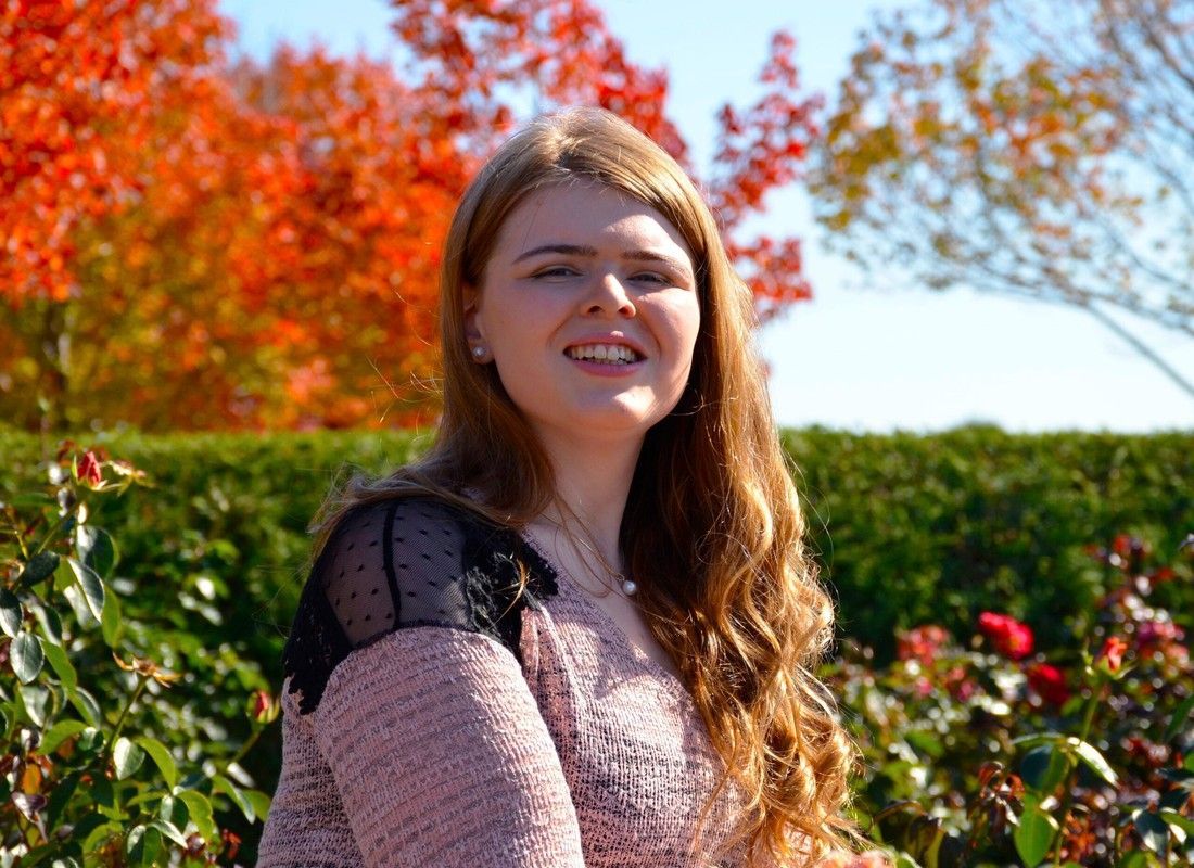 Woman smiling outdoors in a garden with red autumn trees and green hedges in the background