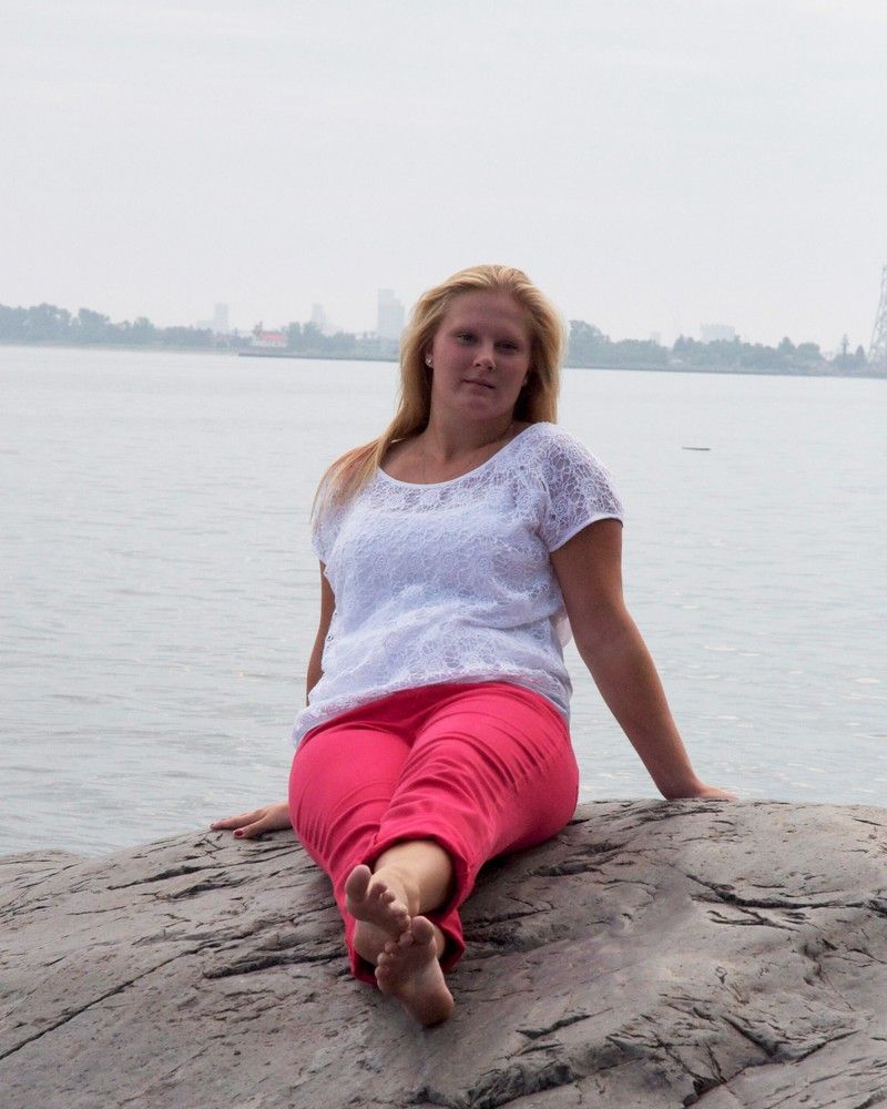 Woman in white top and pink pants sitting on a rock by the water, with a hazy city skyline behind her.
