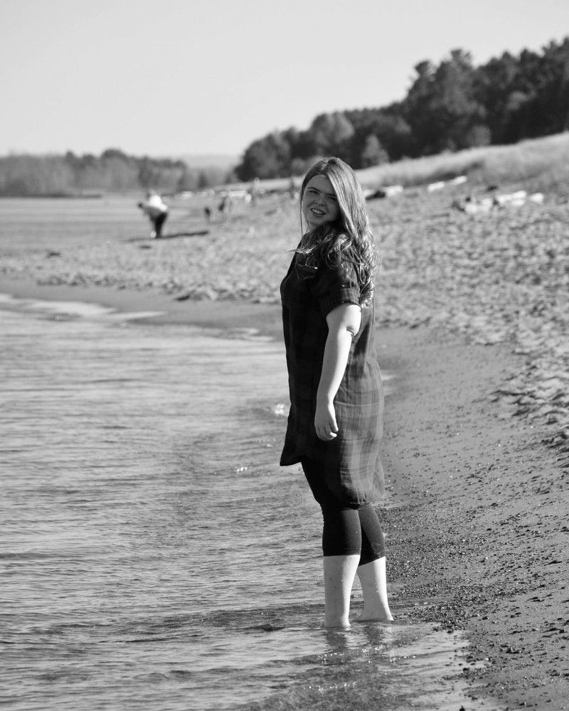 Woman standing in shallow water at a rocky shoreline, facing the camera and smiling.