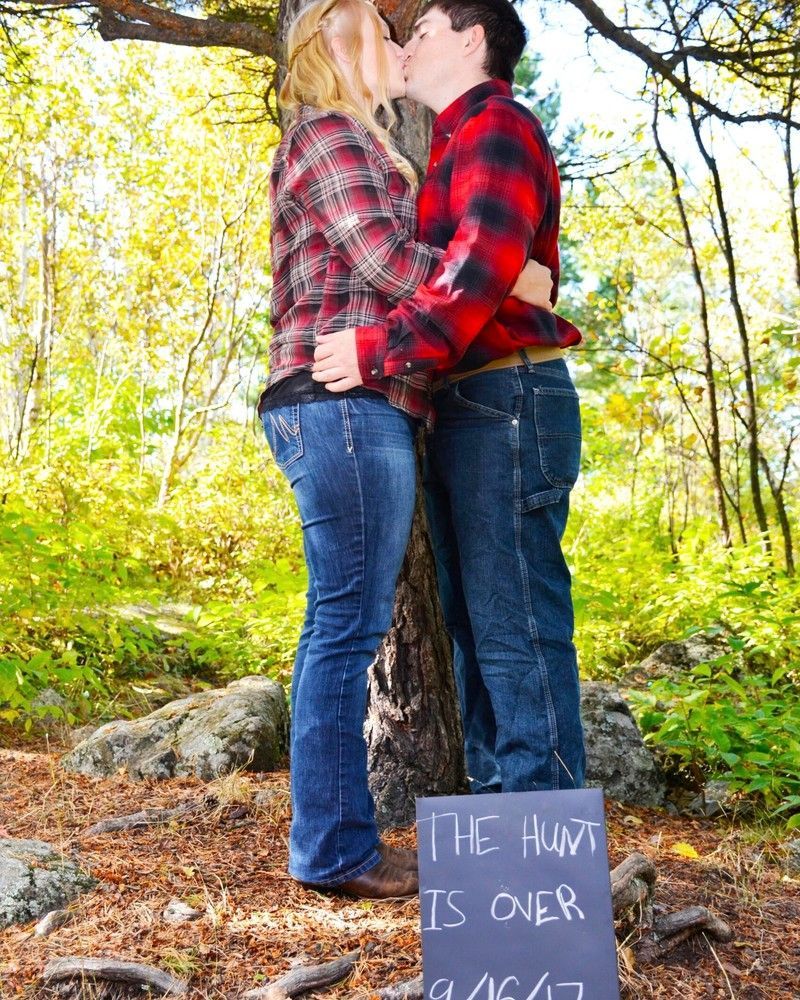 Couple kissing by a tree in a wooded area, with a chalk sign reading “The hunt is over.”