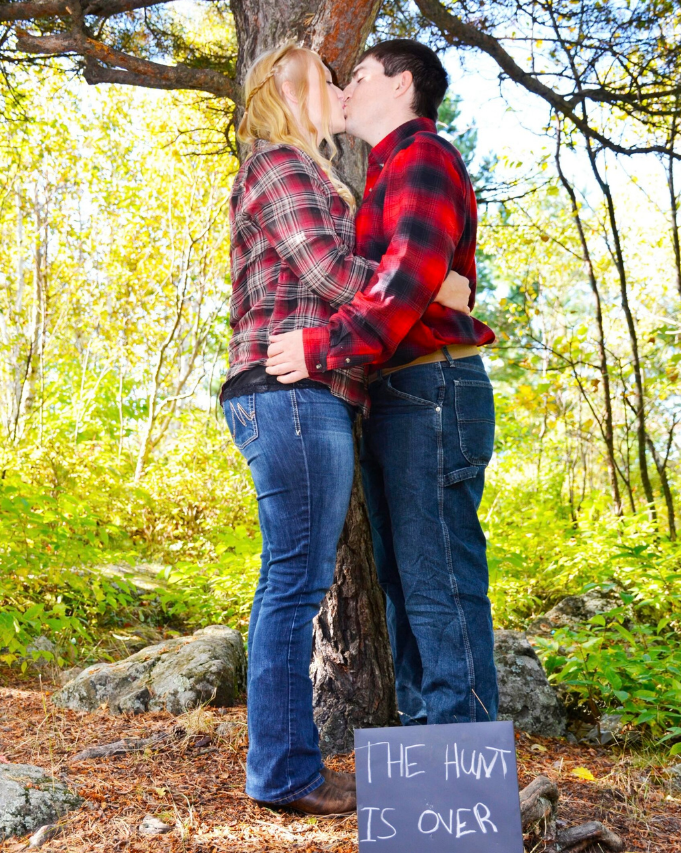 Couple kissing under a tree in a sunny forest, with a sign reading “The hunt is over”