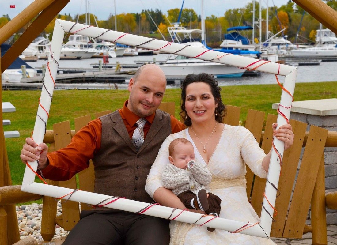 Couple with baby sitting in a decorated wooden frame by a marina, smiling at the camera.