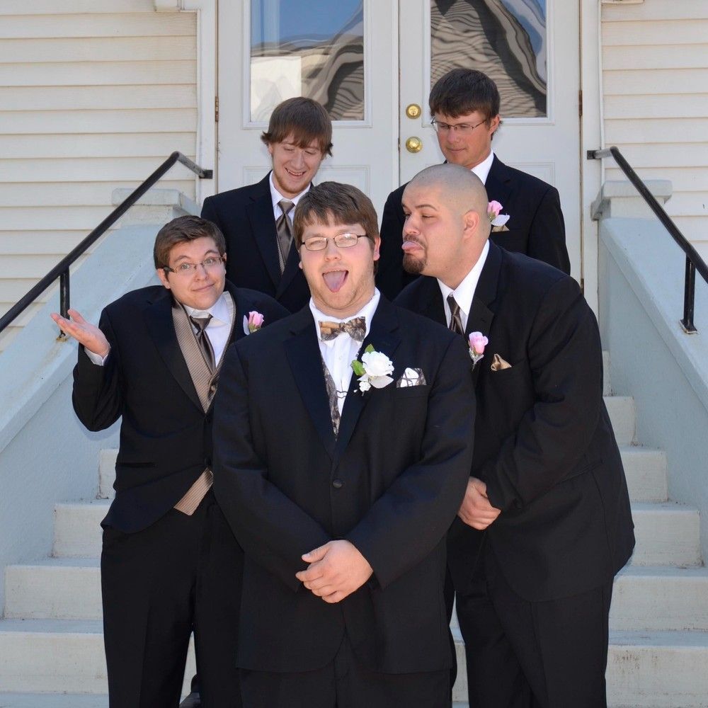 Five people in black suits pose on white steps outside a house, with one making a playful face.