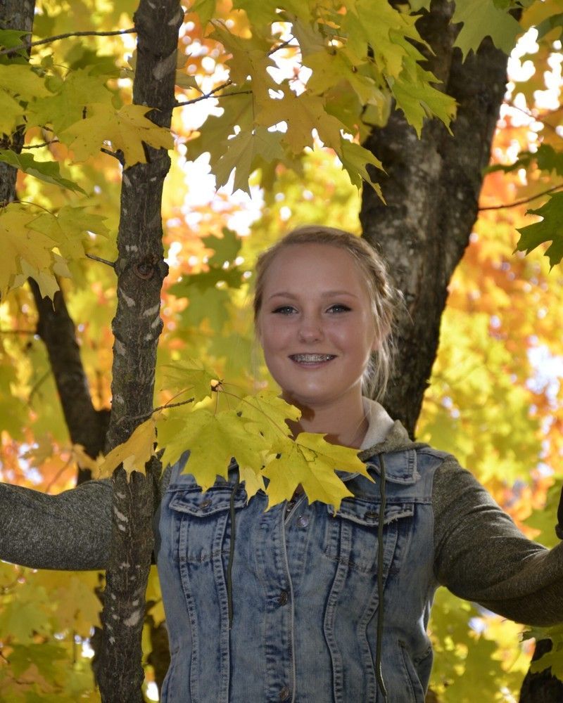 Smiling person in a denim jacket standing among yellow autumn leaves on a tree