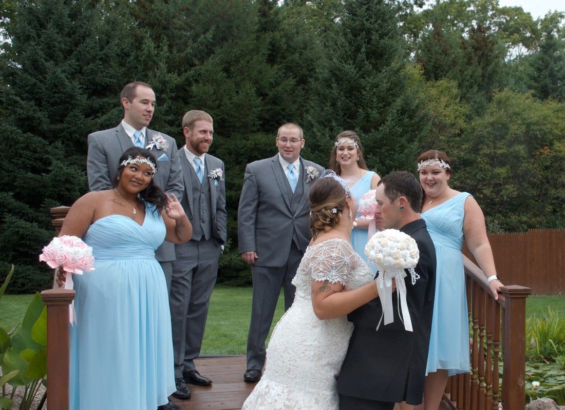 Wedding party posing on a wooden bridge, bride and groom kissing in front, bridesmaids and groomsmen behind.