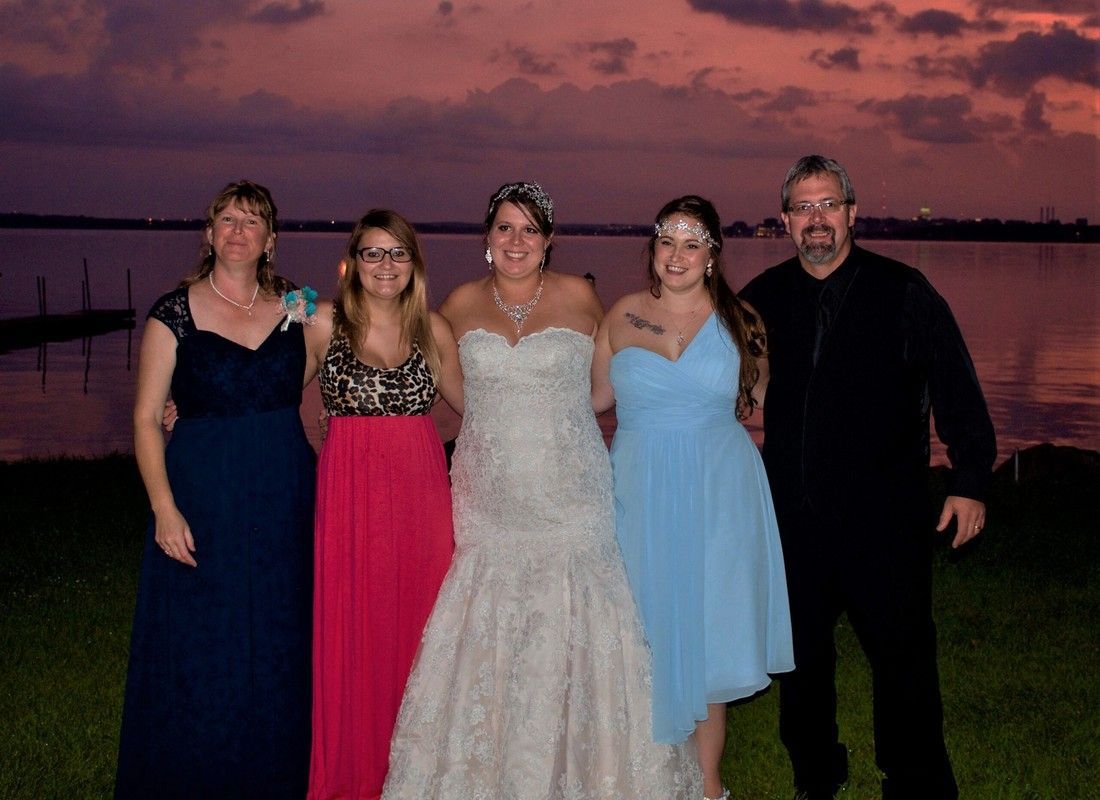 Five people posing in formal attire at sunset near water, with a pink sky behind them