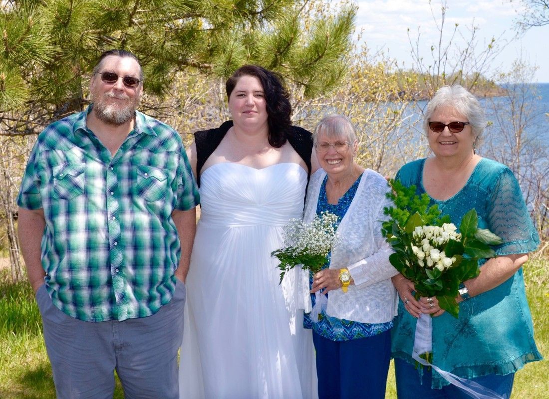 Four people stand outdoors, two holding bouquets, with a woman in a white dress at center.