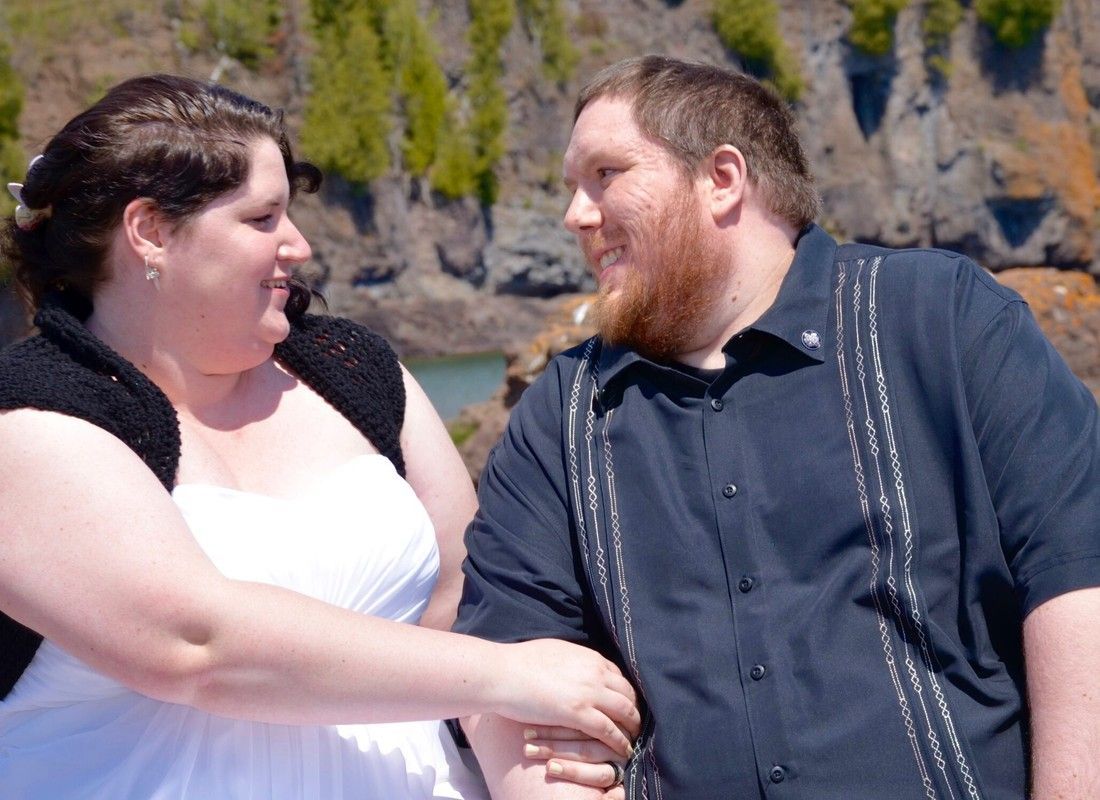 Two people in dark clothing smiling and talking by a rocky waterfront with trees in the background