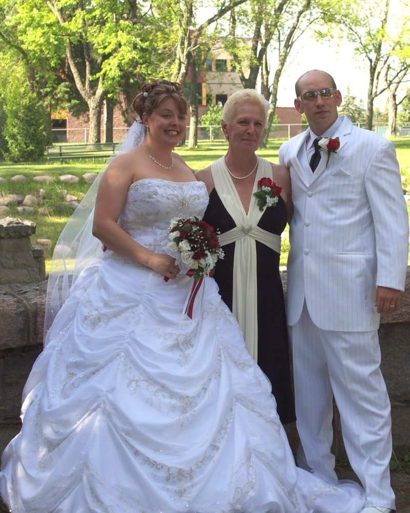 Wedding couple and officiant standing outdoors, bride in white gown and groom in white suit, holding bouquets