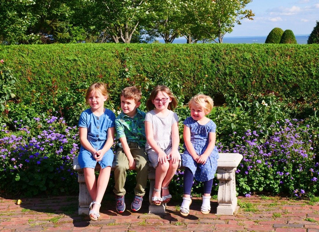 Four children sitting on a bench in a sunny garden with flowers and green hedges