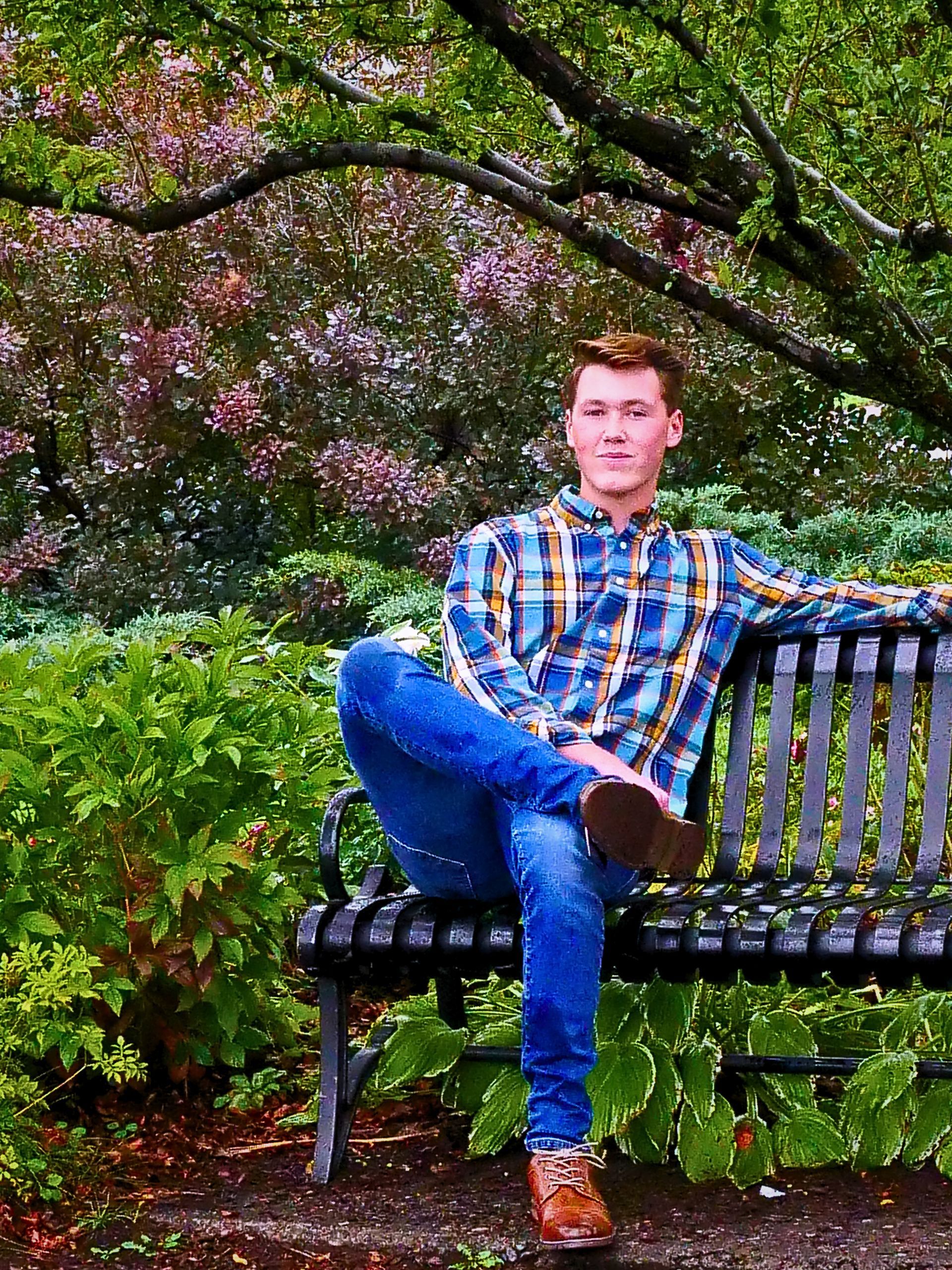 Person sitting cross-legged on a bench in a green garden, wearing a plaid shirt and jeans.