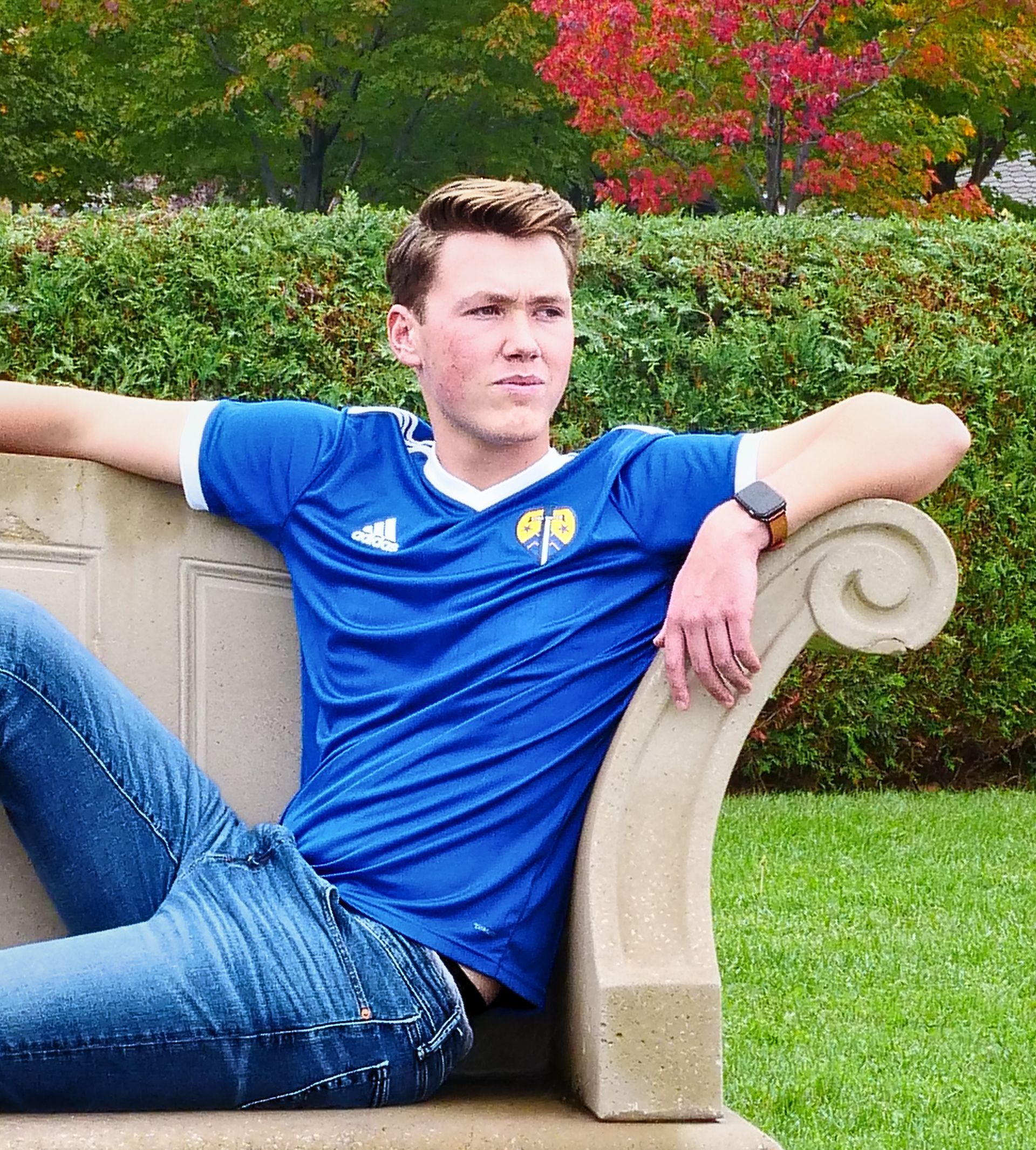 Young man in a blue polo shirt and jeans reclining on a stone bench in a garden.
