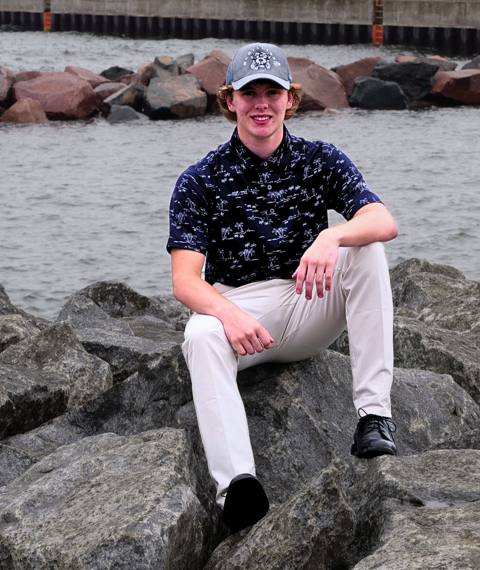 Person sitting on rocks by the water, wearing a black patterned shirt, beige pants, and a cap.