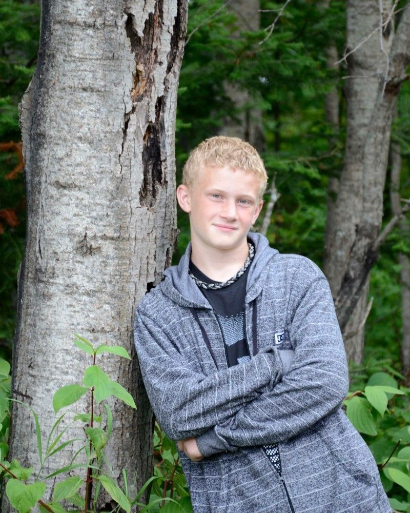Person in a gray jacket leaning against a tree in a wooded area, arms crossed and smiling.