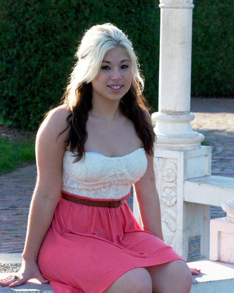 Woman in a pink dress sitting on a white bench outdoors, smiling beside a white column.