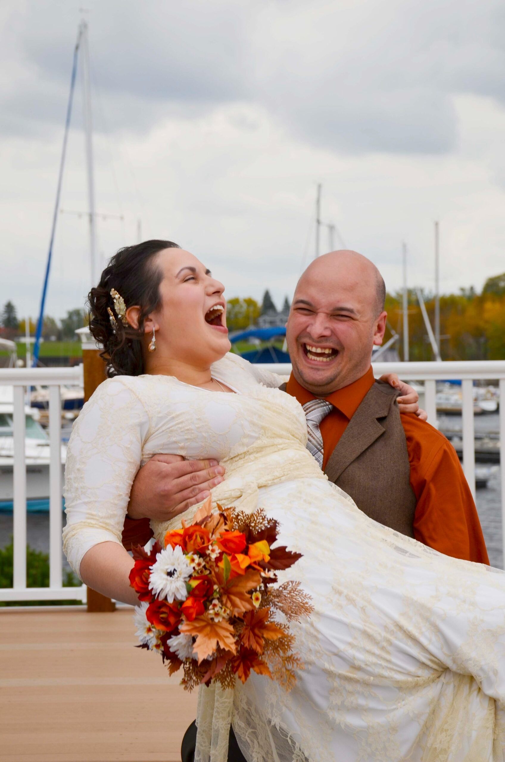 Wedding couple laughing, groom in orange vest carrying bride in white dress and bouquet on a marina dock.