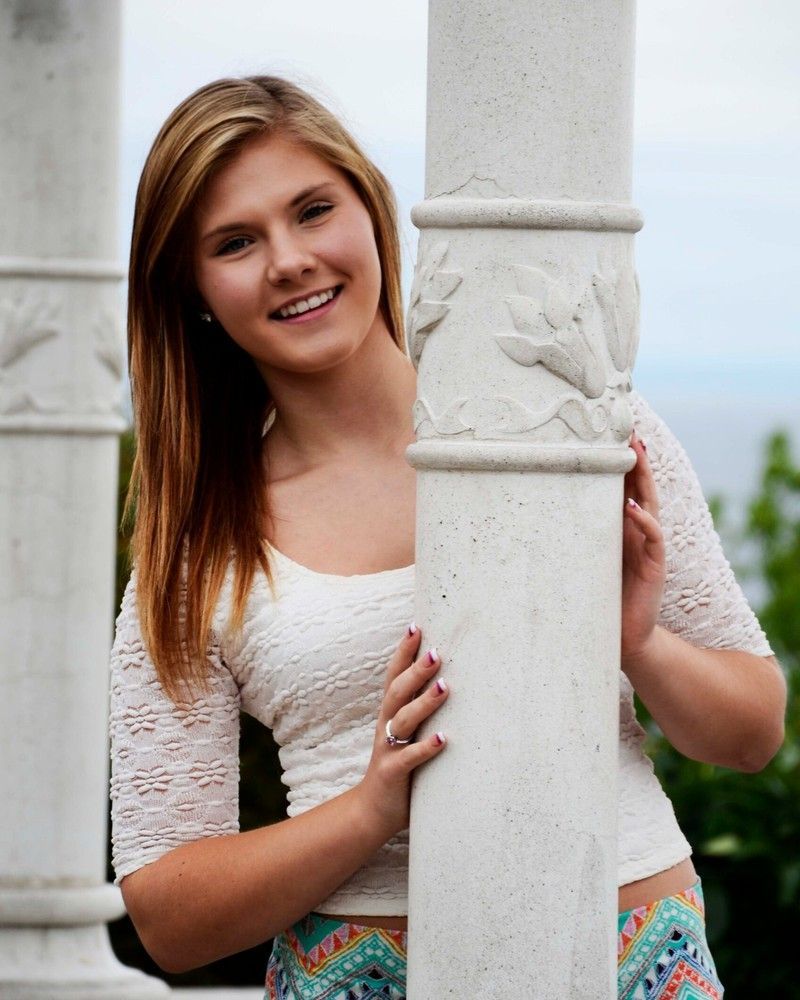 Smiling woman in a white top peeks from behind a white pillar outdoors, wearing a colorful skirt.