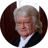 A person with short white hair and glasses wearing a black robe with a white collar, framed in a circular crop.