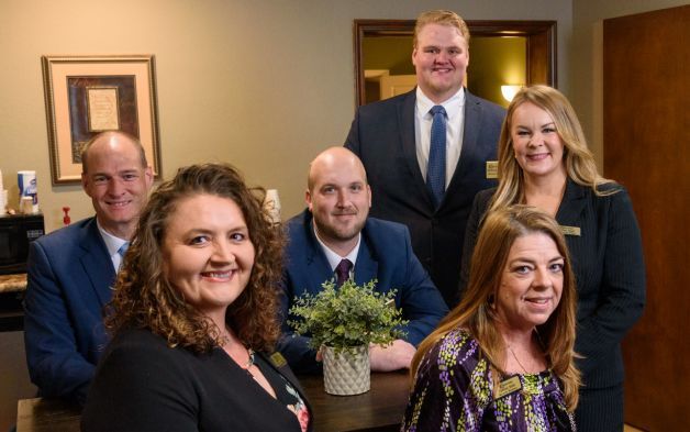 A professional team poses together in an office, with some members standing and others seated behind a table with a plant.