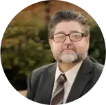 A person with glasses and a goatee, wearing a suit and striped tie, looking directly at the camera.