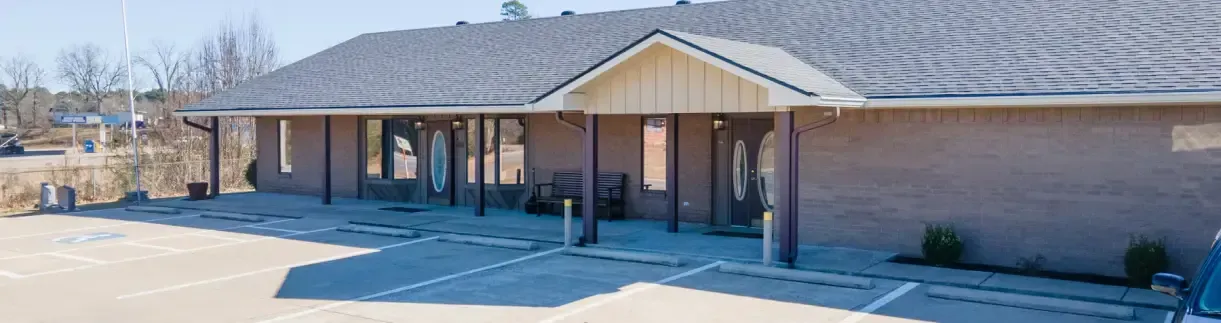 A single-story, tan brick commercial building with a dark shingled roof, a central porch, and a parking lot in front.