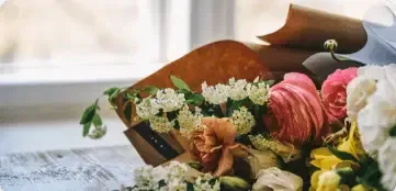 A colorful flower bouquet wrapped in brown paper rests on a light-colored wooden surface near a window.