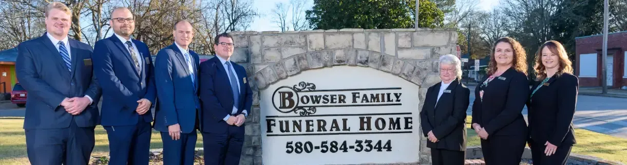 Seven staff members stand in formal attire before a stone sign reading 