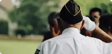A person wearing a white uniform and a military-style garrison cap salutes outdoors, with others blurred in the background.