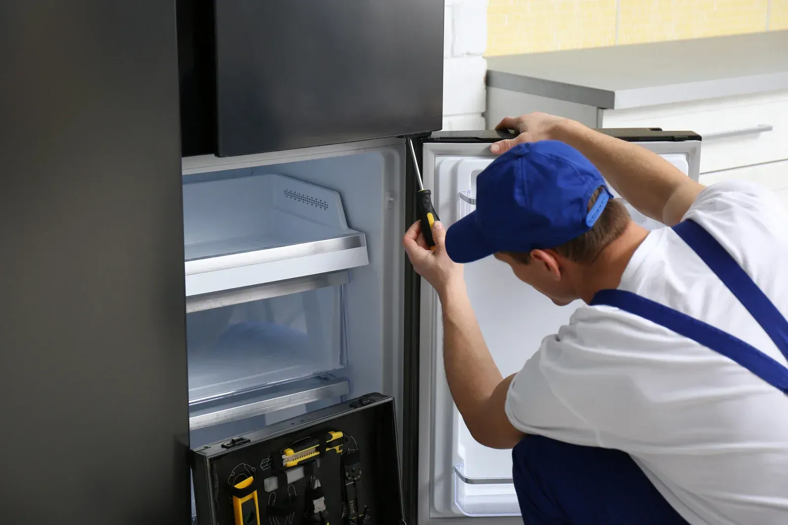 Person in blue cap and overalls repairs a refrigerator door.