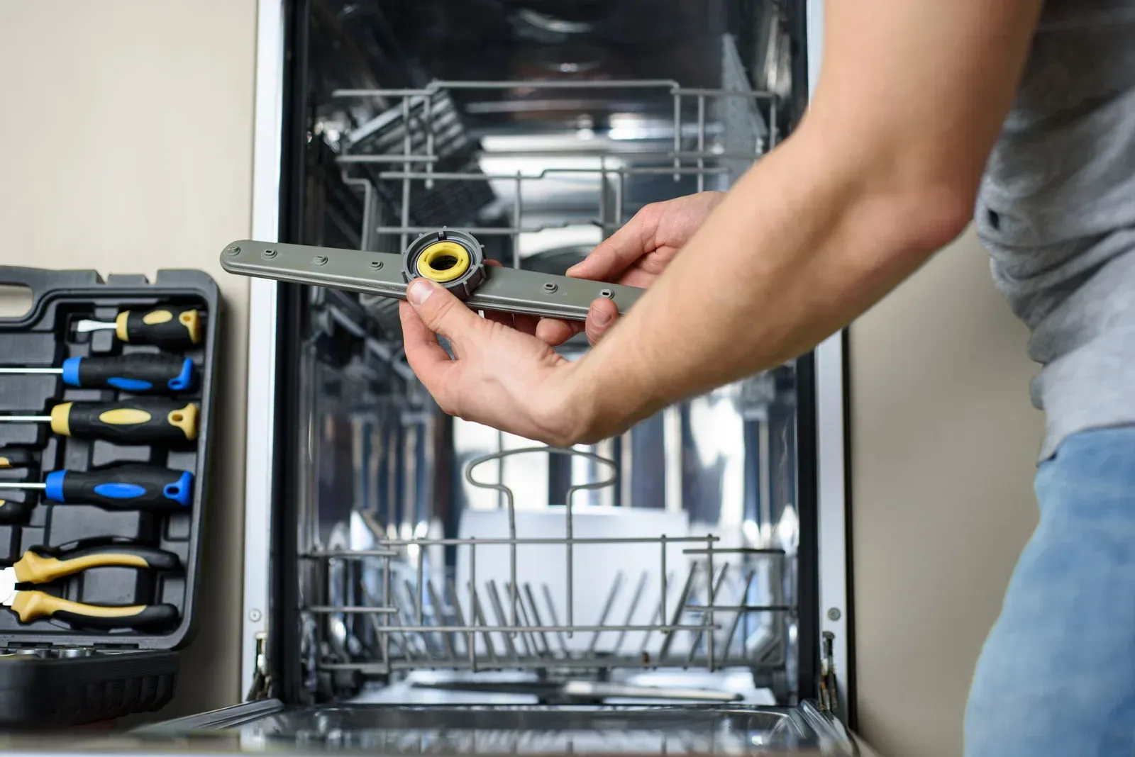Person repairing a dishwasher, holding a sprayer arm. Tools are in a nearby case.