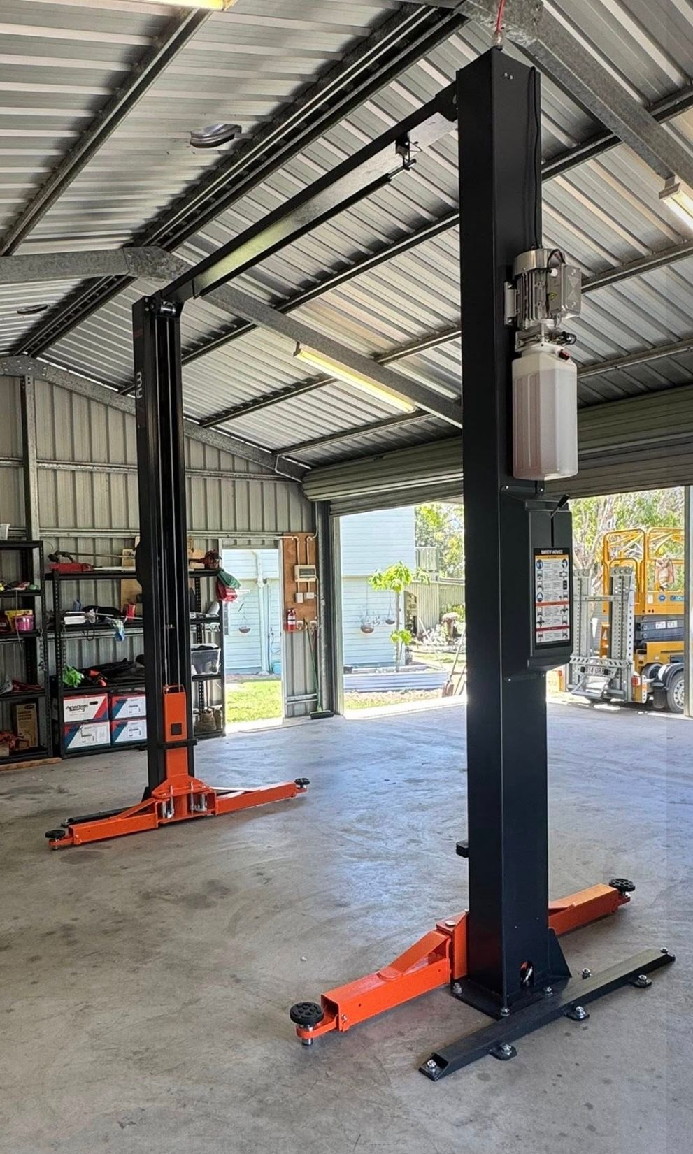 Two-post car lift in a garage with orange base arms and a corrugated metal roof