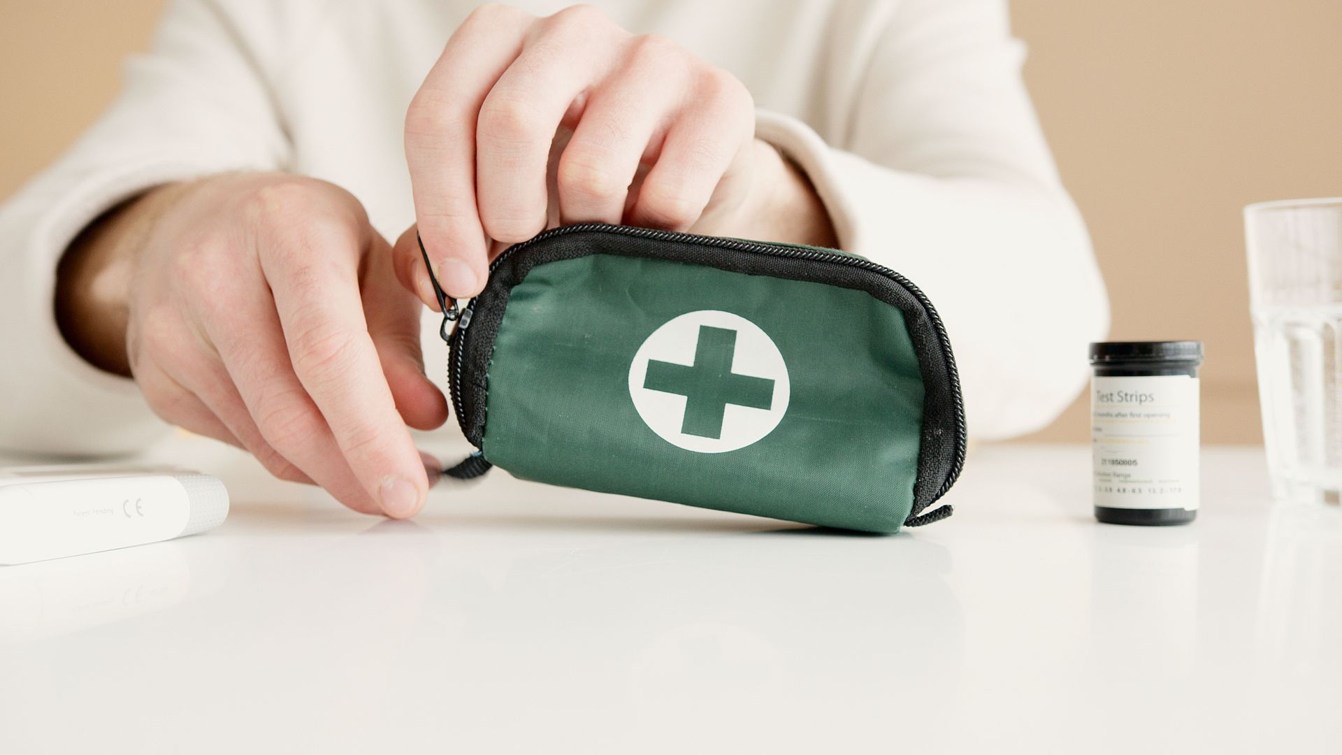 Hands placing a green first-aid kit with a white cross beside a medicine bottle on a white table