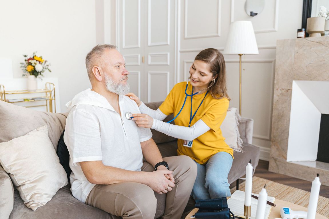Home health worker checking an older man's chest with a stethoscope in a bright living room