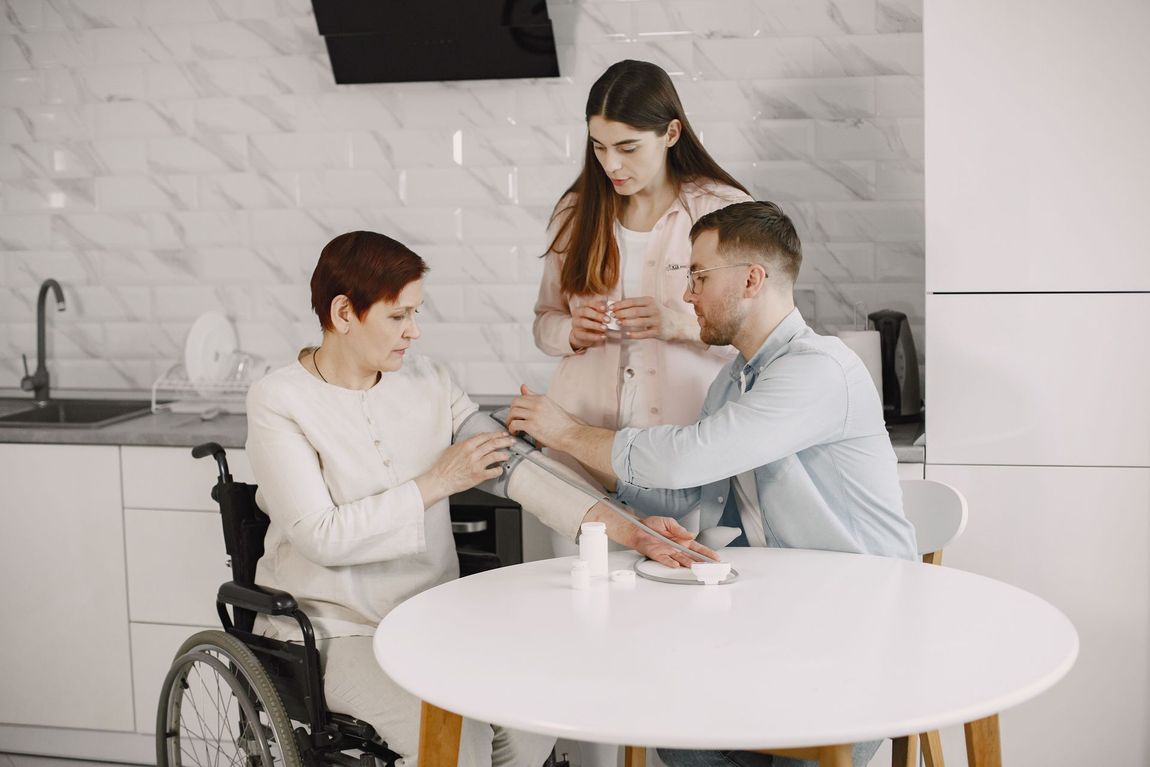 Three people in a kitchen, one seated in a wheelchair, smiling while sharing a phone at a white table