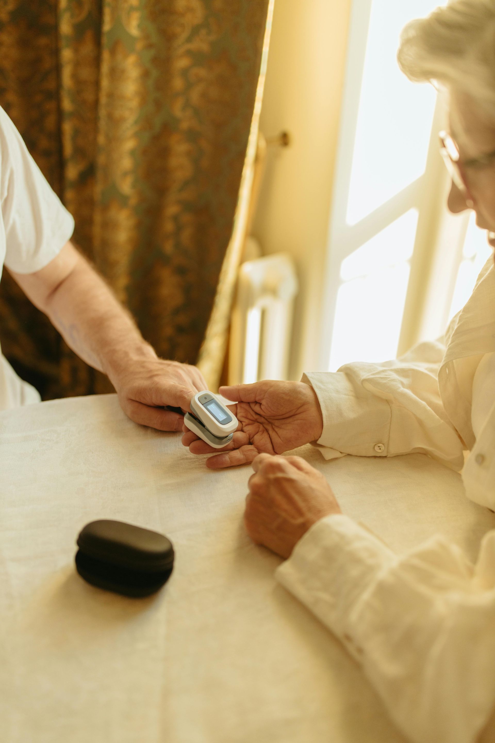 Person checking another person’s finger pulse with a fingertip pulse oximeter at a table by a window