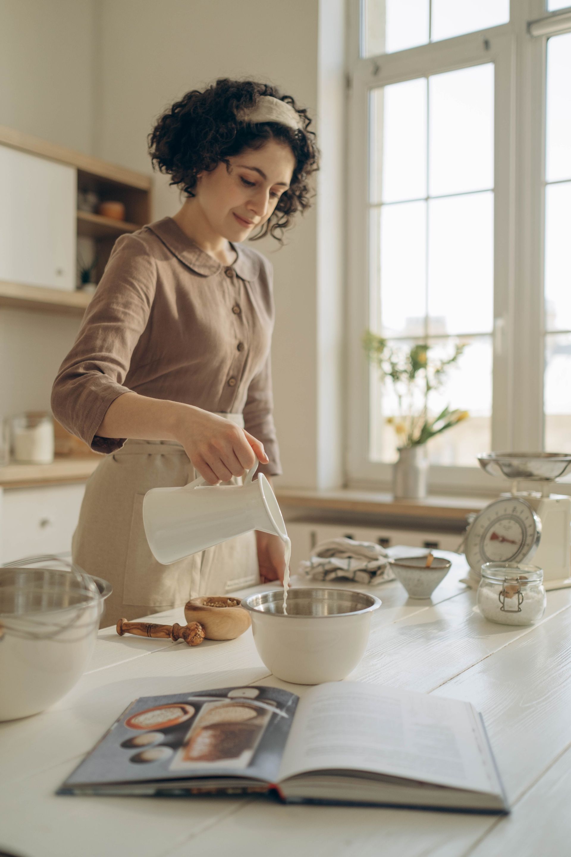 Person pouring batter into a mixing bowl in a bright kitchen, with an open recipe book on the counter.