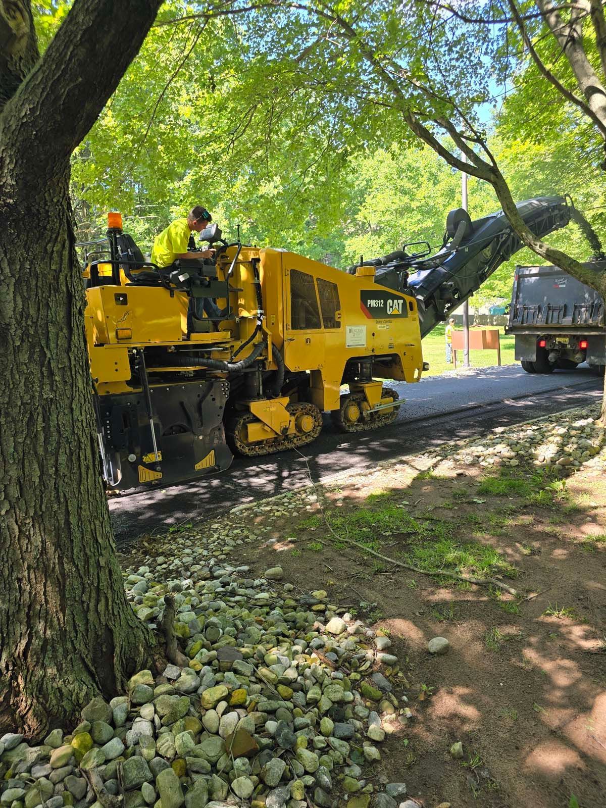A yellow tractor is driving down a road next to a tree.