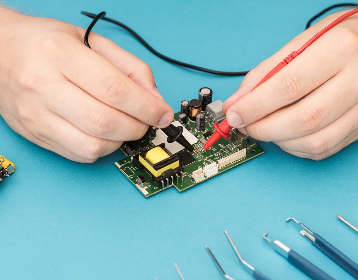 Hands using probes to test a small circuit board on a blue work surface.