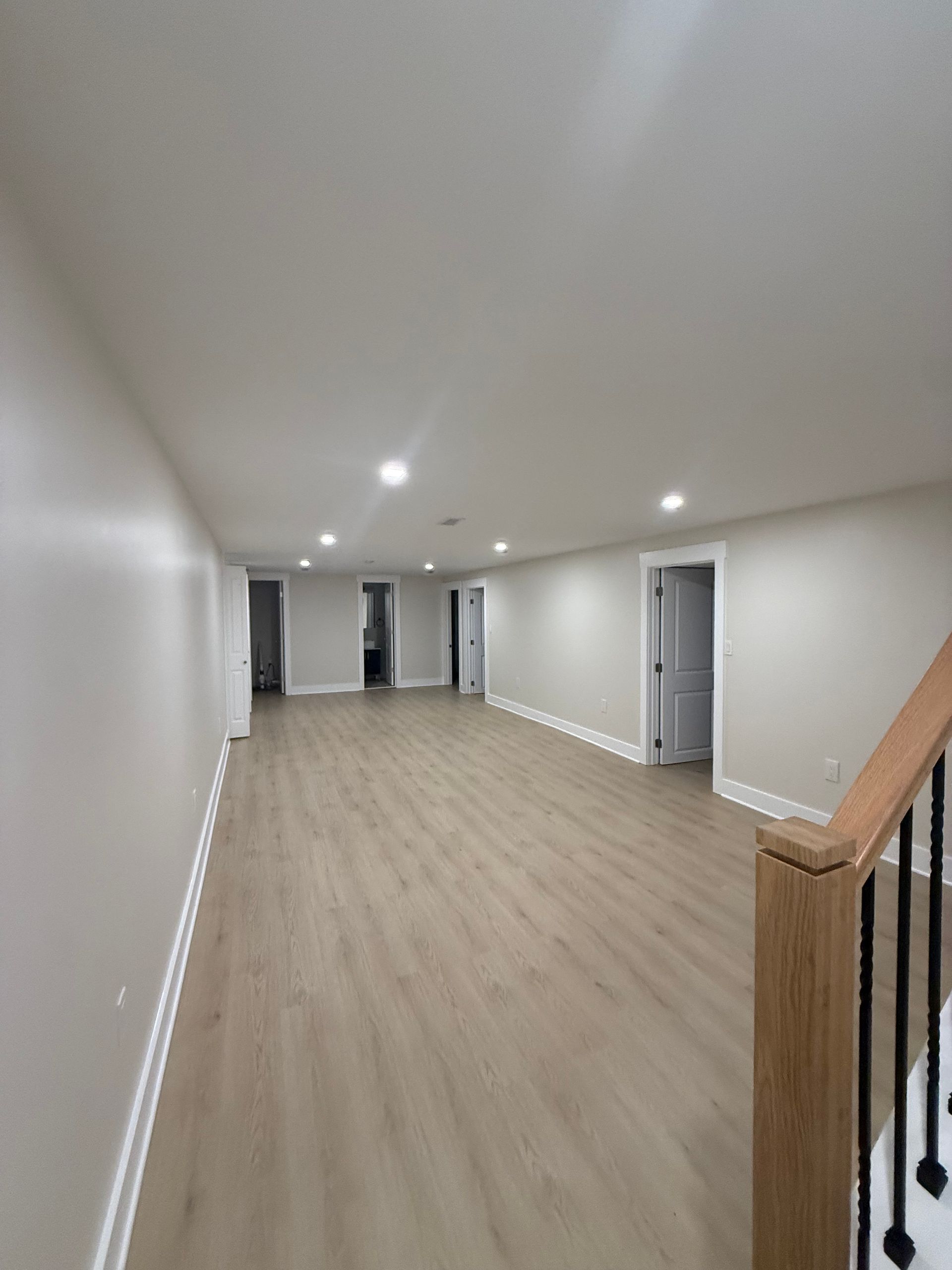 Empty white hallway with wood floors, recessed lights, and a wooden stair railing on the right