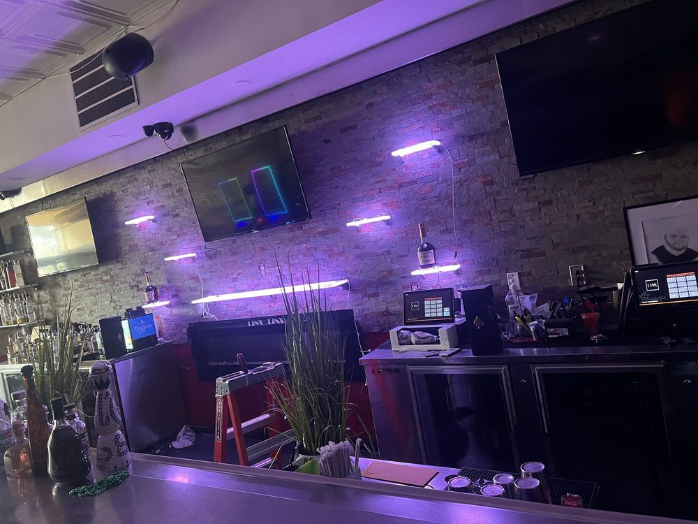 Purple-lit bar counter with brick wall, shelves, and several beer taps under TV screens