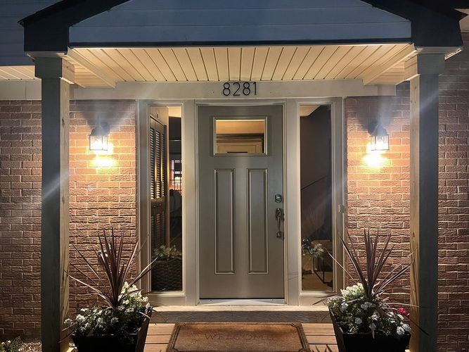 Front entrance of a brick house at dusk, with lit wall lamps and the number 8281 above the door.