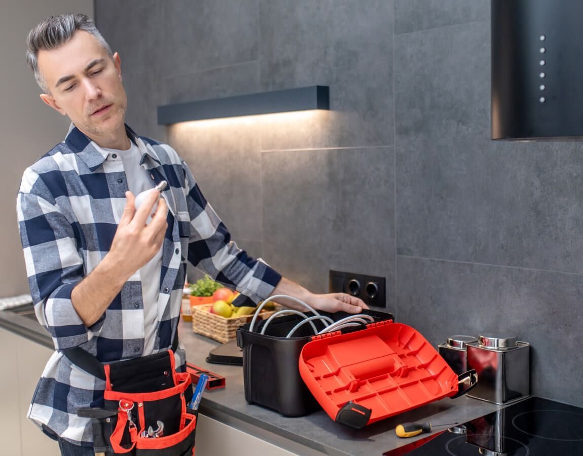 Man in plaid shirt using a tool in a modern kitchen with open tool bag and countertop tools.