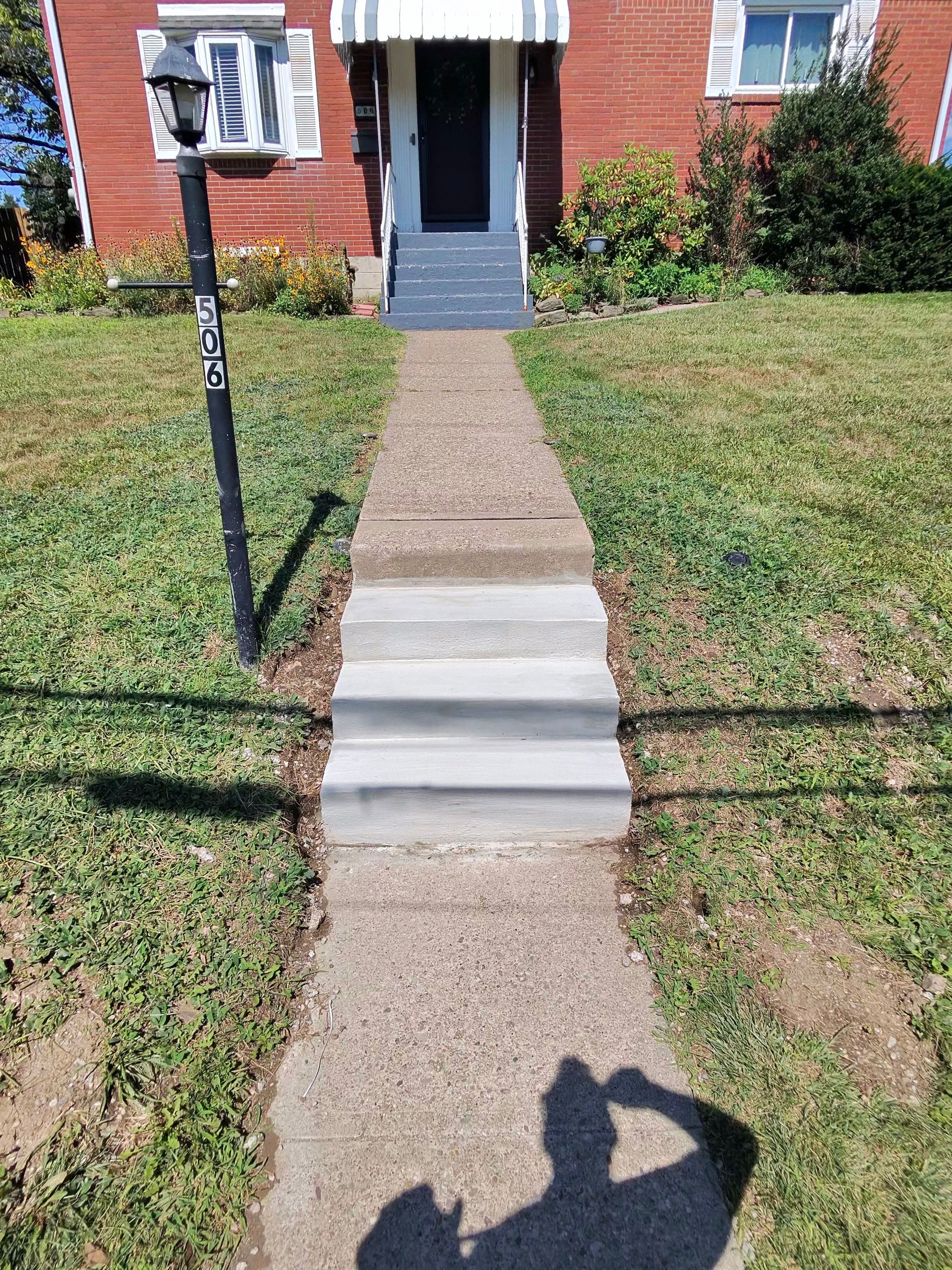 Front walkway with two concrete steps leading to a brick house entrance, bordered by grass and a lamp post.