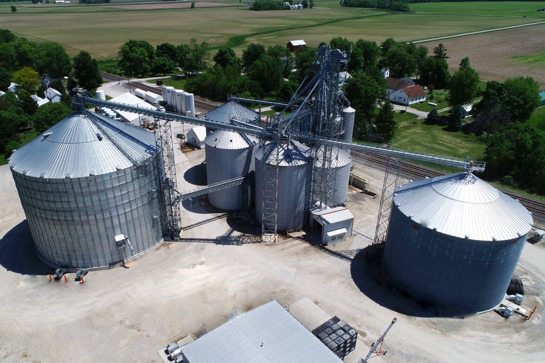 Aerial view of a grain elevator complex with large silver silos and storage bins in a rural field.