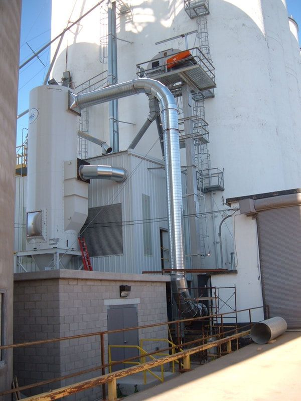 Industrial facility exterior with large silver pipes and white storage tanks beside a brick wall