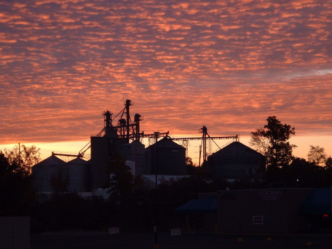 Silhouetted industrial buildings at sunset beneath a dramatic orange and purple cloud-filled sky