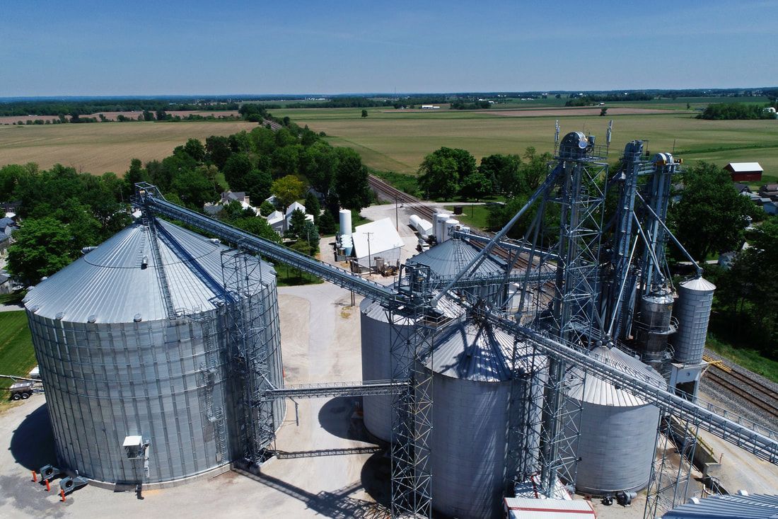 Aerial view of grain silos and conveyor machinery at an agricultural storage facility in open farmland.