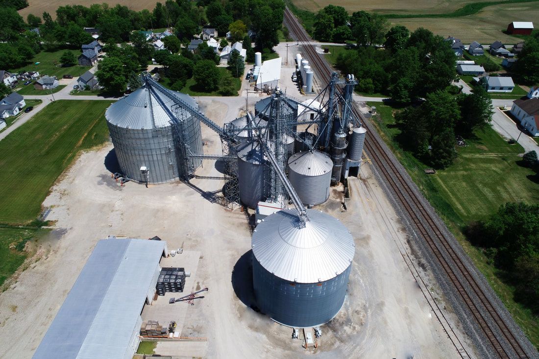 Aerial view of grain silos and industrial storage facility beside railroad tracks and trees.