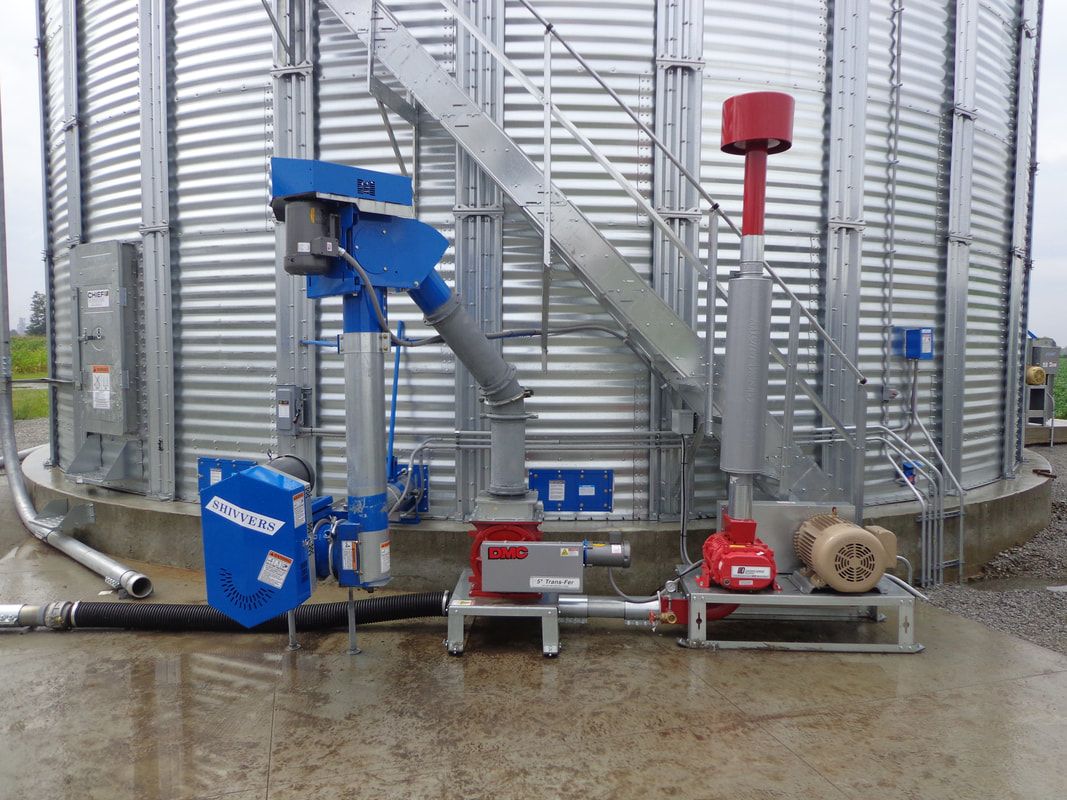 Agricultural grain handling equipment beside large metal silos on a wet concrete pad